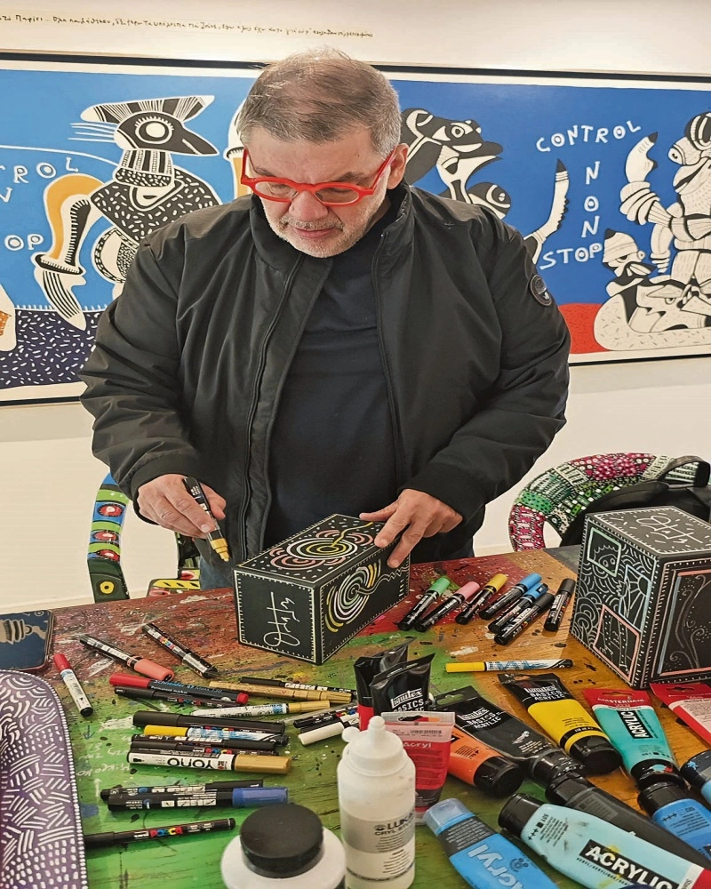 Ζωγραφίζοντας ένα από τα κουτιά που περιέχει τη σειρά έξι βιβλίων του, «Πίνοντας καφέ» με τον Επίκουρο, τον Σωκράτη, τον Αριστοτέλη, τον Θουκυδίδη, τον Πλάτωνα και τον Περικλή.